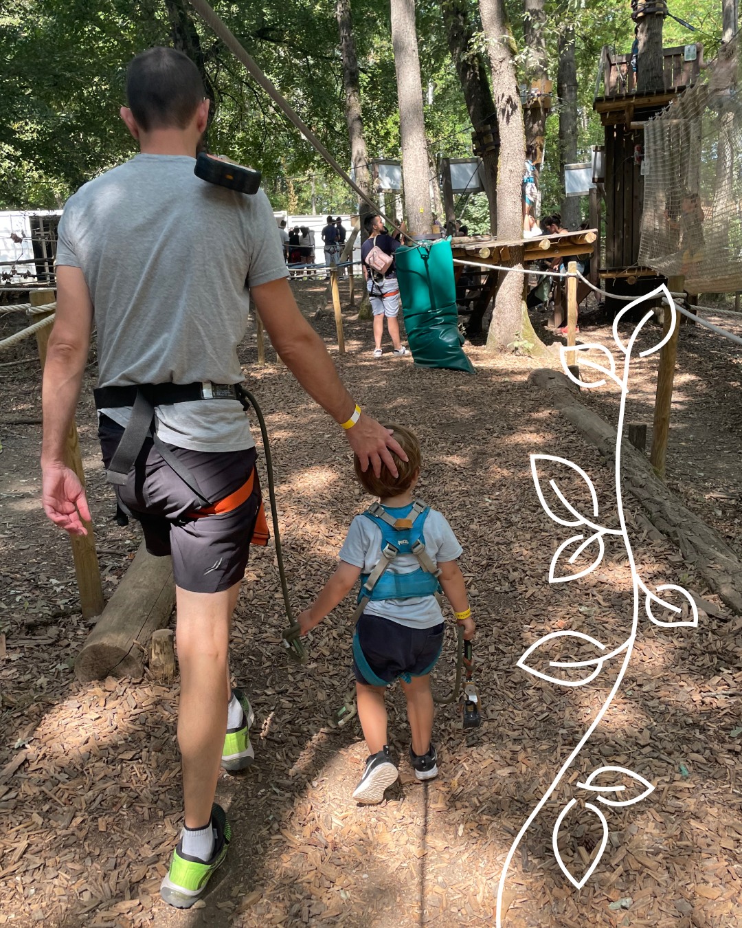 Parcours aventure : adulte et enfant équipés Adulte tenant la main d'un jeune enfant équipé d'un baudrier, marchant sur un sentier forestier vers un parcours aventure.